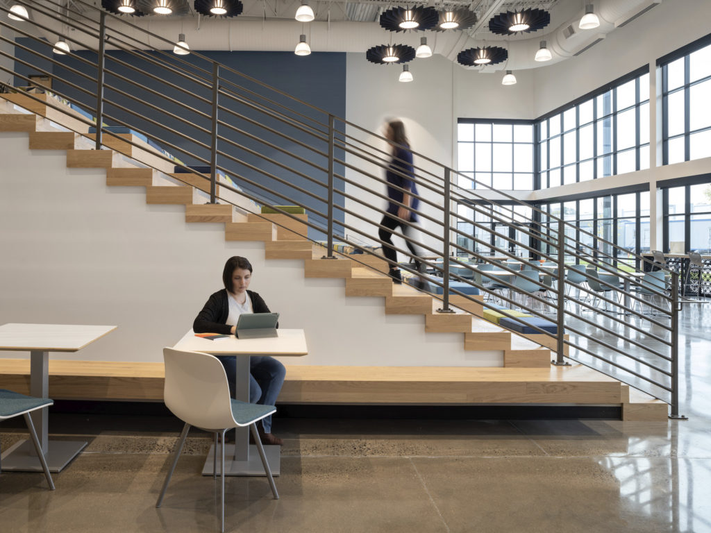 A corporate lobby with a woman sitting at a table and another climbing up beautifully woodworked stairs.