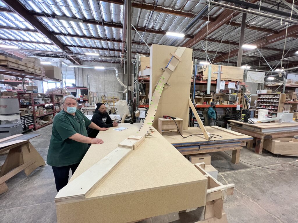 Two workers in the Faustrollean woodshop, one man in a green shirt and mask, the other in a hoodie, are standing by a large workstation with various tools and materials in the background.